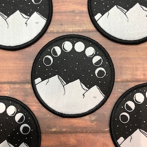 May include: A set of four black and white embroidered patches featuring a mountain range with a crescent moon phase arc above. The patches are circular and have a white outline.