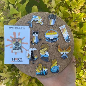 May include: A selection of enamel pins with diverse designs, such as a cat, fish, and phoenix, arranged on a corkboard. A card with the text "HARTIFUL.CO.UK" and a logo is also visible. The pins are vibrant and eye-catching.