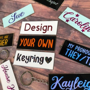 Puede incluir: Una colección de llaveros coloridos con diferentes diseños y textos. Los llaveros presentan nombres, frases como "Design Your Own" y "Keyring", y pronombres. Los llaveros están hechos de metal y tienen un acabado plateado.