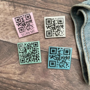May include: Four square-shaped enamel pins with QR codes. One is pink with "SCAN ME" text, another is white, and the other two are teal and green. The pins are on a wooden surface.