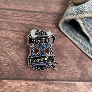 May include: A silver-colored enamel pin featuring an hourglass design with the words "Time To Procrastinate." The pin has a blue and silver color scheme with a crescent moon and star at the top. The pin is on a wooden surface.