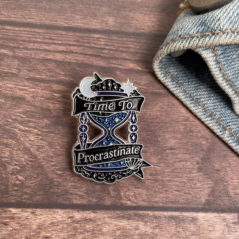 May include: A silver-colored enamel pin featuring an hourglass design with the words "Time To Procrastinate." The pin has a blue and silver color scheme with a crescent moon and star at the top. The pin is on a wooden surface.