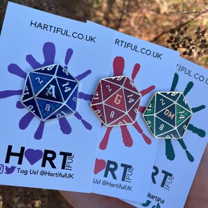 May include: Three enamel pins shaped like 20-sided dice with different colors and glitter. The dice are blue, red, and green. The blue die has the letter A on it, the red die has the letter G on it, and the green die has the letters DM on it.