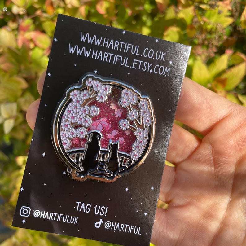 Puede incluir: Un pin redondo de esmalte plateado con la silueta de un perro y un gato en un puente bajo un cerezo en flor. El fondo es rosa con purpurina. El pin est&aacute; sobre una tarjeta negra con el texto "www.hartiful.co.uk" y "www.hartiful.etsy.com".