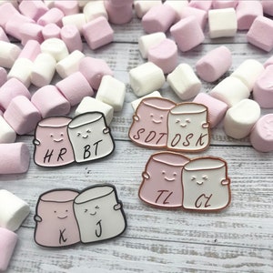 May include: Three pairs of pink and white enamel pins shaped like marshmallows with smiling faces. Each pair has a different set of initials: HR & BT, SDT & OSK, and TL & CJ.
