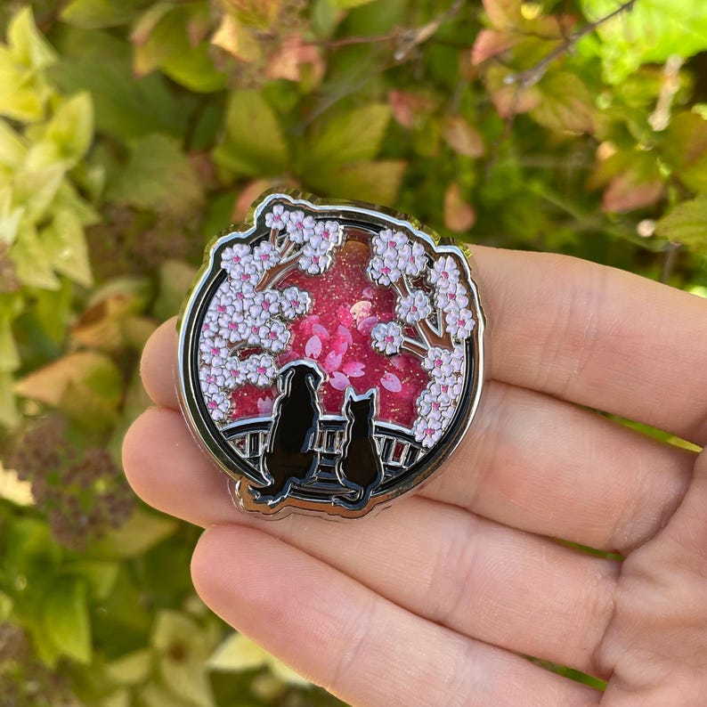 Puede incluir: Un pin redondo de esmalte plateado que representa la silueta negra de un perro y un gato mirando una escena de flores de cerezo. El fondo es de un rosa vibrante con flores blancas y rosas. El pin se sostiene en una mano.