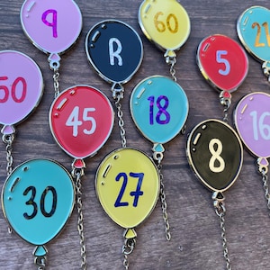 Könnte beinhalten: Eine Sammlung bunter Emaille-Anhänger in Ballonform, die jeweils mit einer individuellen Zahl oder einem Buchstaben versehen sind. Die Ballons sind in Rosa, Rot, Gelb, Schwarz und Türkis gehalten, mit silbernen Umrandungen und Metallketten.