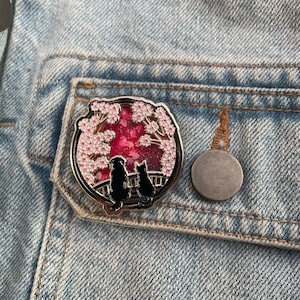 Puede incluir: Un pin redondo de esmalte plateado que representa la silueta de un perro y un gato bajo un cerezo en flor. El fondo es rojo vibrante con detalles rosas. El pin est&aacute; colocado sobre una chaqueta vaquera azul claro.