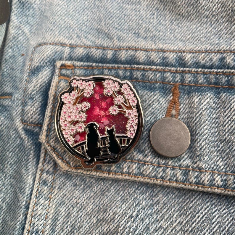 Puede incluir: Un pin redondo de esmalte plateado que representa la silueta de un perro y un gato bajo un cerezo en flor. El fondo es rojo vibrante con detalles rosas. El pin est&aacute; colocado sobre una chaqueta vaquera azul claro.