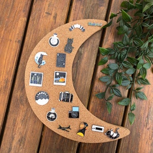 May include: A corkboard shaped like a crescent moon with various enamel pins attached. The pins feature images of a cat, a suitcase, a camera, a plane, a bottle, a moon, and a building. The corkboard is on a wooden surface.