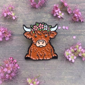 May include: A brown Highland cow enamel pin with a floral crown of pink and white flowers. The pin is on a wooden background.