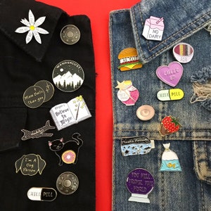 May include: Two denim jackets with a variety of enamel pins attached. The pins feature various designs, including a cat with a donut, a book with the words "Believe in Magic", a plane, a dog, a pill, a mountain range, a fairy dust jar, a no dairy sign, a burger, a heart with the words "Cheer Up", a pill with the words "Chill Pill", a fish in a bag, a globe with the words "You Will Travel the World", and a strawberry.