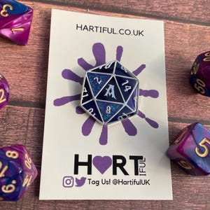 May include: A silver and blue enamel pin shaped like a 20-sided die with glitter accents. The die has the letter "A" in the center and the numbers 1, 2, 4, 8, 14, 20 around the edges. The pin is on a white card with the text "HARTIFUL.CO.UK" and "HORT" with a purple heart.
