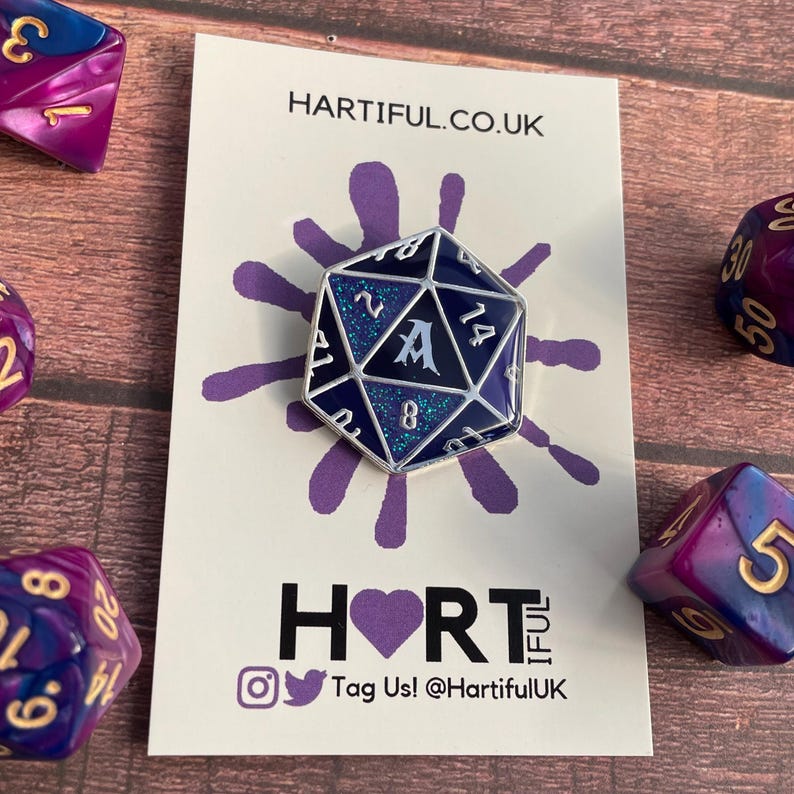 May include: A silver and blue enamel pin shaped like a 20-sided die with glitter accents. The die has the letter "A" in the center and the numbers 1, 2, 4, 8, 14, 20 around the edges. The pin is on a white card with the text "HARTIFUL.CO.UK" and "HORT" with a purple heart.