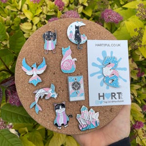 May include: A collection of enamel pins on a corkboard, featuring various designs in pastel colors. Designs include a dachshund, cats, a phoenix, a fish, a tarot card, crystals, and a dragon, all with transgender pride flag colors. A card with the text "HARTIFUL.CO.UK" is also present.