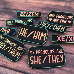 Peut inclure: Un ensemble de neuf &eacute;pingles en &eacute;mail noir avec des bordures arc-en-ciel iris&eacute;es. Chaque &eacute;pingle pr&eacute;sente un ensemble de pronoms diff&eacute;rent, notamment "ZE/ZEM", "ANY PRONOUNS ARE FINE", "THEY/THEM", "HE/HIM", "XE/XE", "MY PRONOUNS ARE THEY/THEM", "MY PRONOUNS ARE SHE/THEY", et "ANY PRONOUNS".