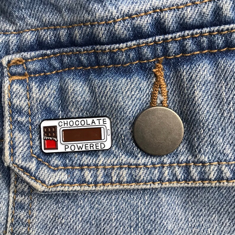 K&ouml;nnte beinhalten: Eine wei&szlig;e Emaille-Anstecknadel mit einer Schokoladentafel-Grafik und dem Text "Chocolate Powered". Die Anstecknadel ist an einer Jeansjacke befestigt.