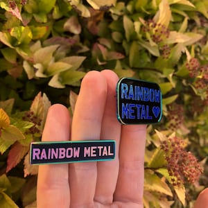 Peut inclure: Deux &eacute;pingles en m&eacute;tal arc-en-ciel avec le texte "Rainbow Metal" dessus. Une &eacute;pingle est noire avec un c&oelig;ur bleu et l'autre est noire avec un c&oelig;ur rose.