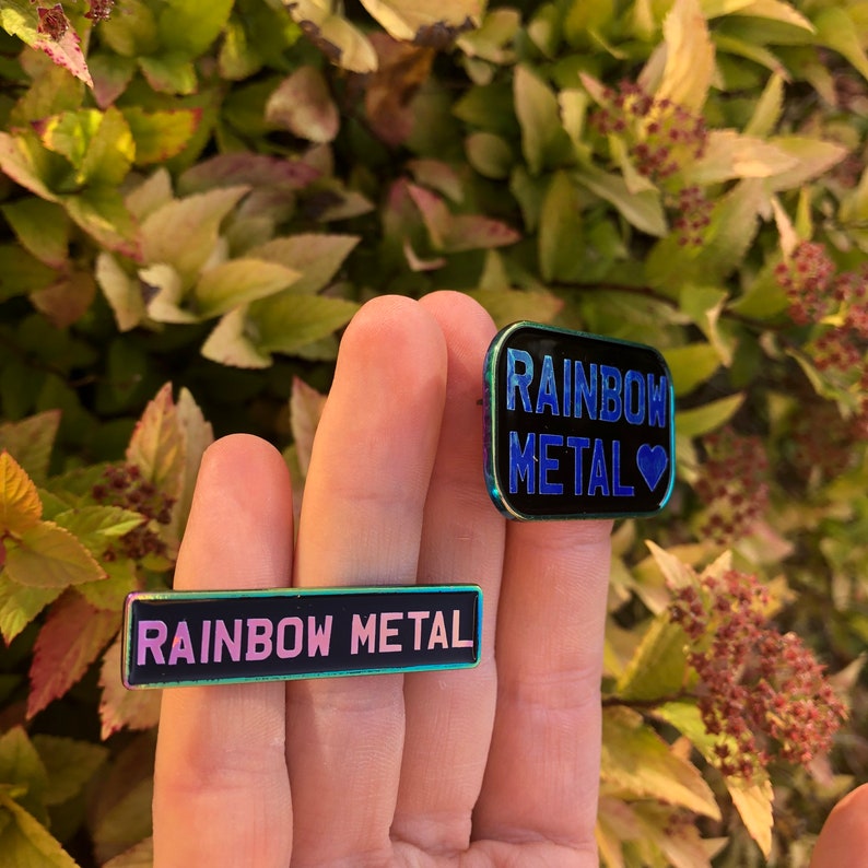 Peut inclure: Deux &eacute;pingles en m&eacute;tal arc-en-ciel avec le texte "Rainbow Metal" dessus. Une &eacute;pingle est noire avec un c&oelig;ur bleu et l'autre est noire avec un c&oelig;ur rose.