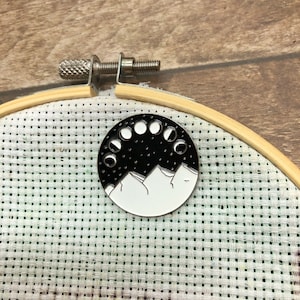 May include: A white enamel pin with a black and white design of a mountain range with a crescent moon phase cycle above it.
