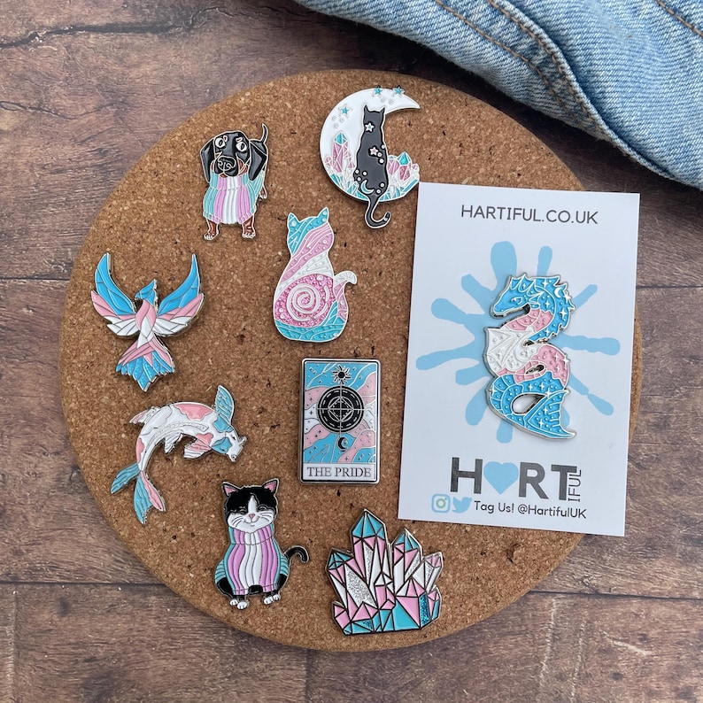 May include: A collection of enamel pins on a corkboard, featuring various designs such as a dachshund, cats, a phoenix, a fish, a tarot card, and crystals. The pins incorporate pink, blue, and white colors, with the text "THE PRIDE" visible on one.