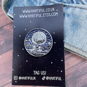May include: A round enamel pin featuring a whale tail design against a full moon and starry night sky. The pin has a blue and silver color scheme. The card behind the pin has the website address www.hartiful.co.uk and www.hartiful.etsy.com.
