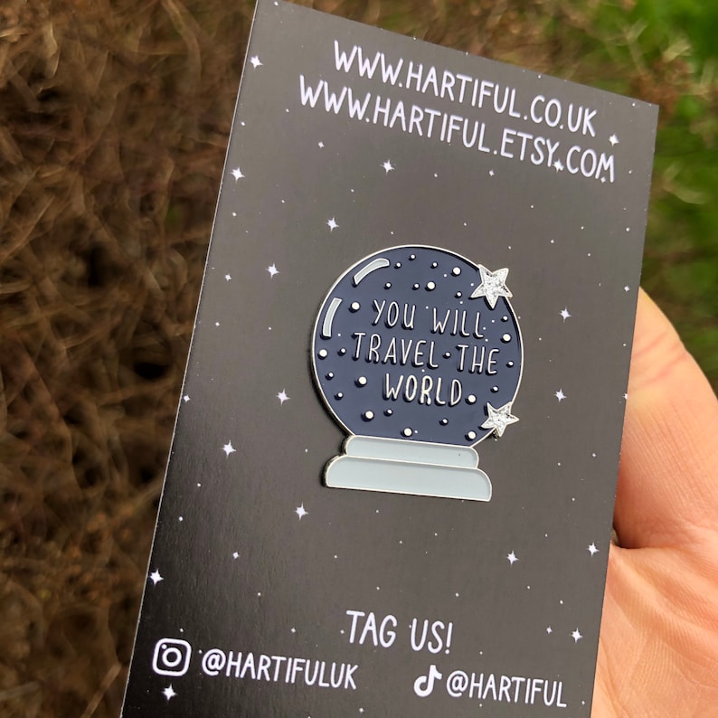 K&ouml;nnte beinhalten: Eine schwarz-wei&szlig;e Emaille-Anstecknadel mit einem blauen Glasglobus mit dem Text "You will travel the world" und wei&szlig;en Sternen. Die Anstecknadel befindet sich auf einem schwarzen Hintergrund mit wei&szlig;em Text, der "www.hartiful.co.uk" und "www.hartiful.etsy.com" lautet.