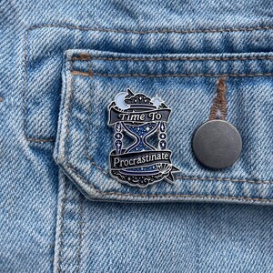 May include: A black and silver enamel pin with the words "Time To Procrastinate" on a denim jacket. The pin features an hourglass design with a crescent moon and stars at the top. A round, gray button is visible.
