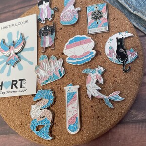 May include: A collection of enamel pins with a transgender pride theme. The pins feature various designs, including animals, crystals, and a tarot card, in shades of pink, blue, and white. The pins are displayed on a corkboard.