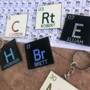 May include: A set of six keychains featuring a black and white periodic table element design. Each keychain has a different element symbol and name, including Carbon, Hydrogen, Bromine,  and Elijah. The keychains are made of metal and have a silver finish.