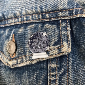 K&ouml;nnte beinhalten: Eine schwarze und wei&szlig;e Emaille-Anstecknadel in Form einer Kristallkugel mit dem Text "You will travel the world" im Inneren. Die Anstecknadel hat eine silberne Oberfl&auml;che und ist an einer Jeansjacke befestigt.
