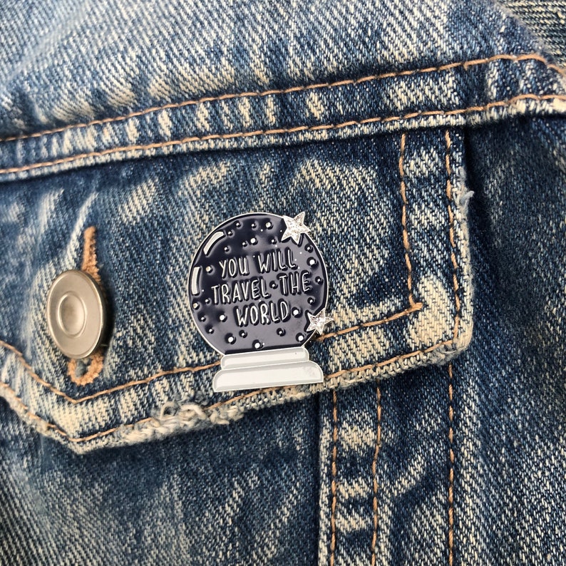 K&ouml;nnte beinhalten: Eine schwarze und wei&szlig;e Emaille-Anstecknadel in Form einer Kristallkugel mit dem Text "You will travel the world" im Inneren. Die Anstecknadel hat eine silberne Oberfl&auml;che und ist an einer Jeansjacke befestigt.