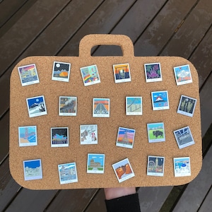 May include: A corkboard with 25 colorful enamel pins featuring various cityscapes and landmarks. The pins are arranged in a grid pattern on a brown corkboard.
