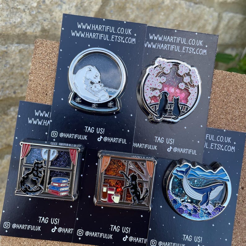 Puede incluir: Una colecci&oacute;n de pines esmaltados con varios dise&ntilde;os. Un pin representa un oso polar y su cachorro en una bola de nieve. Otros pines muestran gatos, flores de cerezo y una ballena. Cada pin est&aacute; en una tarjeta negra con informaci&oacute;n de sitio web y redes sociales.