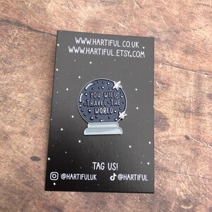 K&ouml;nnte beinhalten: Eine schwarz-wei&szlig;e Emaille-Anstecknadel mit einem blauen Glasglobus mit dem Text "You will travel the world" und Sternen. Die Anstecknadel befindet sich auf einem schwarzen Hintergrund mit der Website-Adresse www.hartiful.co.uk und www.hartiful.etsy.com. Der Text "Tag us!" befindet sich unter der Anstecknadel mit den Social-Media-Handles @hartifuluk und @hartiful.