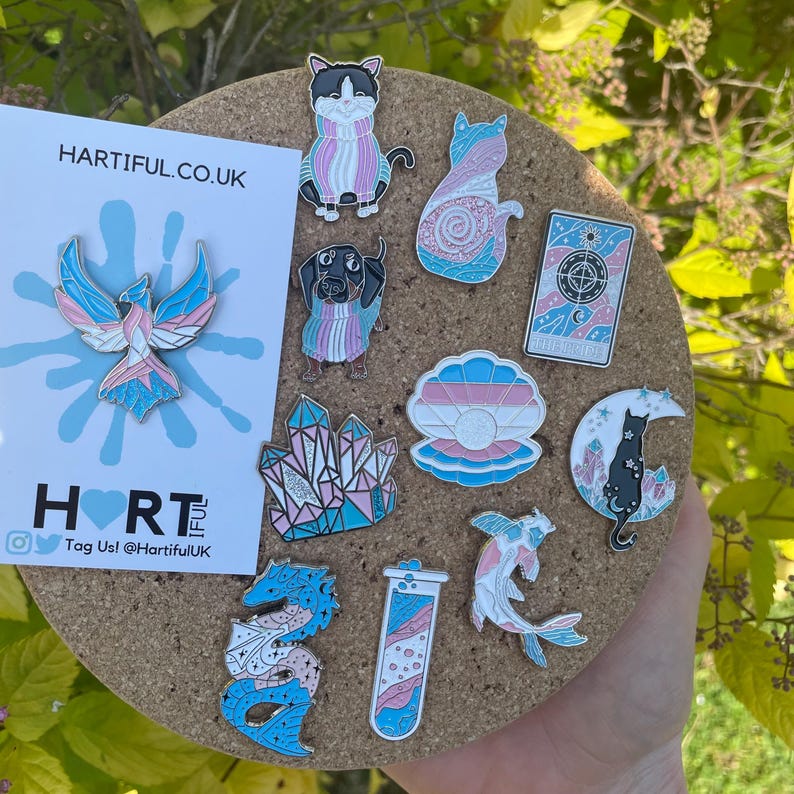 May include: A collection of enamel pins on a corkboard, featuring various designs in blue, pink, and white. Designs include a phoenix, cats, a dachshund, crystals, a seashell, a tarot card, a cat on a crescent moon, a dragon, a test tube, and a koi fish. The text "HARTIFUL.CO.UK" is visible.
