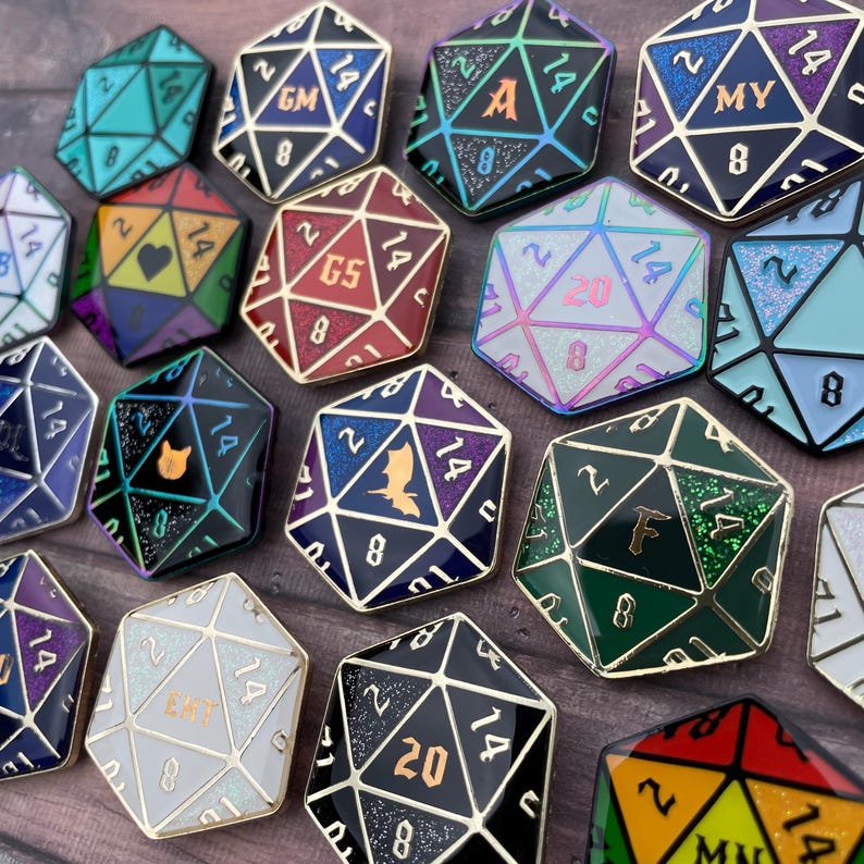 May include: A collection of 12 enamel pins shaped like 20-sided dice. Each die has a different color and design, including a black die with a bat, a green die with a letter F, and a white die with the letters "EMT".