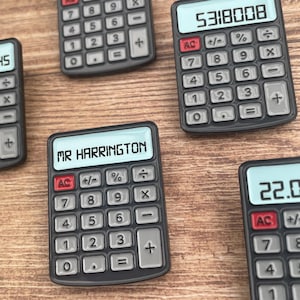 May include: Several small, dark gray calculators with white number displays are arranged on a wooden surface. One calculator displays "MR HARRINGTON" on its screen. Other calculators show numerical values. The calculators have a standard button layout.