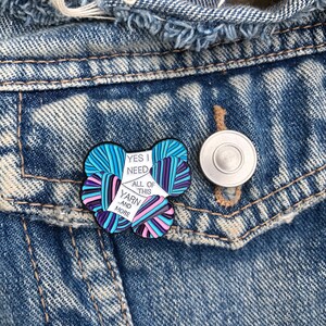 May include: A white enamel pin with a colorful yarn design. The pin says "Yes I need all of this yarn and more".