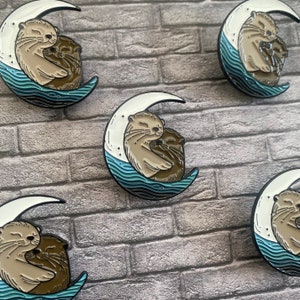 May include: Enamel pins featuring two otters cuddling on a crescent moon. The moon is white with a black outline, and the water is blue. The otters are light and dark brown. The pins are set against a gray brick background.