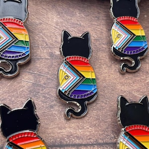 May include: Black cat enamel pins with rainbow and transgender pride flag designs. The pins feature a black cat silhouette wearing a sweater with the colors of the pride flag. The pins are set against a brown wooden surface.