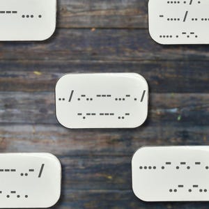 May include: White rectangular magnets with Morse code patterns. The magnets are arranged on a wooden surface. The Morse code includes dots, dashes, and spaces, forming a message. The background is a rustic wooden texture.