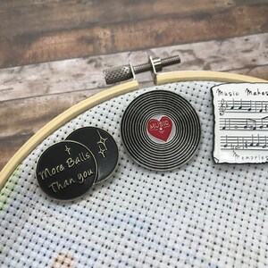 May include: Three enamel pins featuring a black and gold pin with the text "More Balls Than You", a black and silver pin with a red heart and the text "Music" inside, and a black and white pin with musical notes and the text "Music Makes Memories".