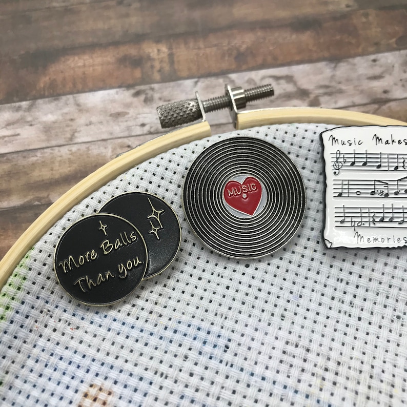 May include: Three enamel pins featuring a black and gold pin with the text "More Balls Than You", a black and silver pin with a red heart and the text "Music" inside, and a black and white pin with musical notes and the text "Music Makes Memories".
