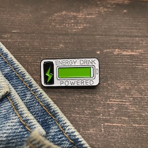 May include: A rectangular enamel pin with a white border, featuring the words "ENERGY DRINK POWERED". The pin has a green battery level indicator and a black energy drink can with a green lightning bolt.