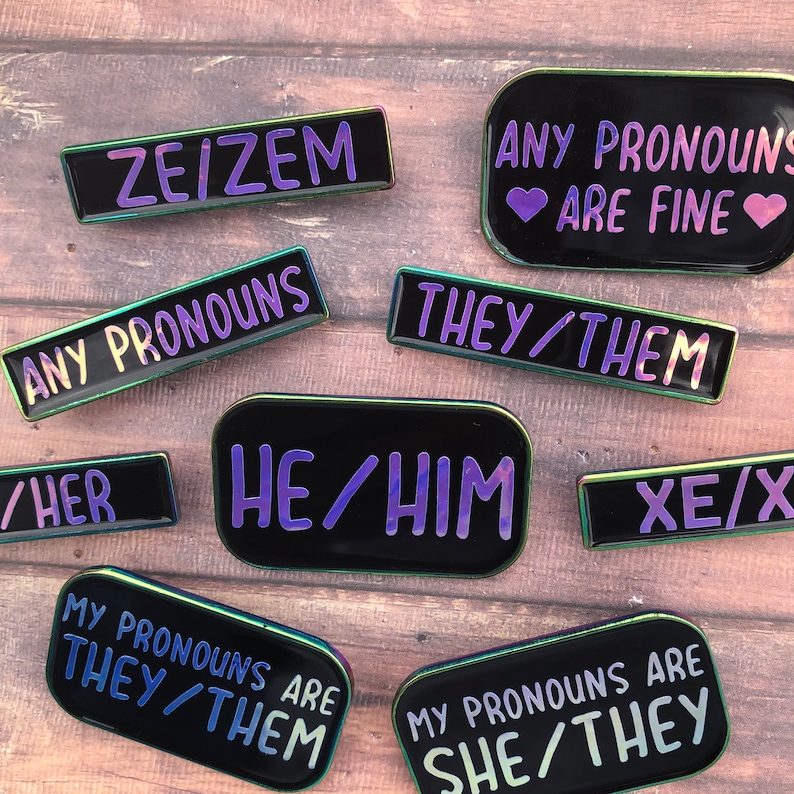 Peut inclure: Un ensemble de dix &eacute;pingles en &eacute;mail avec diff&eacute;rentes options de pronoms, notamment "ZE/ZEM", "ANY PRONOUNS ARE FINE", "THEY/THEM", "HE/HIM", "XE/X", "MY PRONOUNS ARE THEY/THEM", et "MY PRONOUNS ARE SHE/THEY". Les &eacute;pingles sont noires avec des accents arc-en-ciel iris&eacute;s.