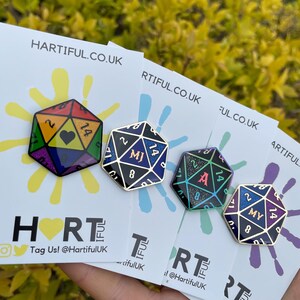 May include: Four enamel pins shaped like a 20-sided die with different colored backgrounds and glitter accents. The pins feature the initials "MJ", "A", and "MY" in gold lettering. The pins are displayed on white cards with the text "Hartiful.co.uk" printed on them.