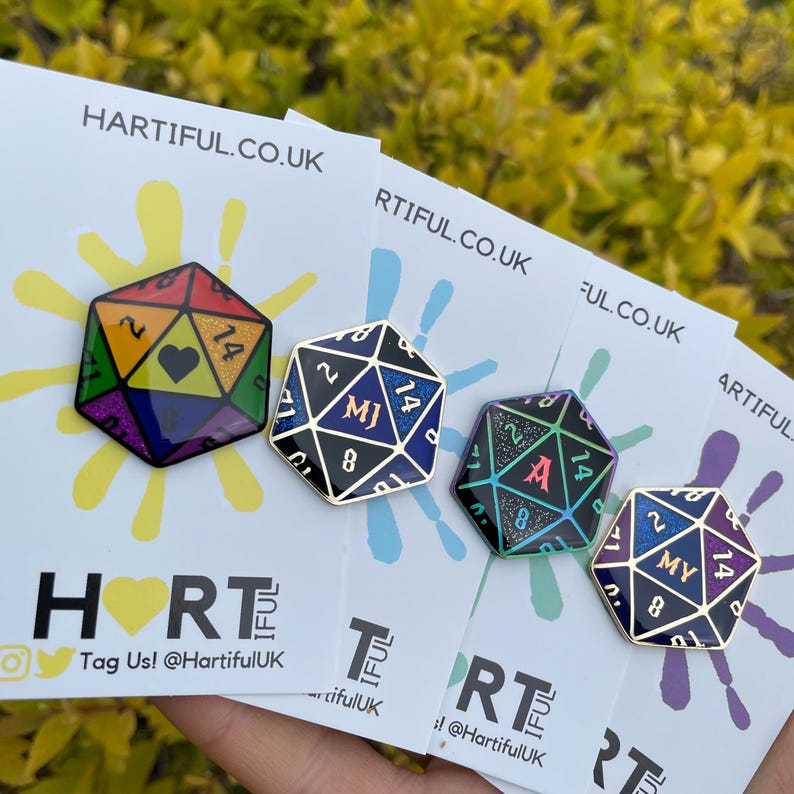 May include: Four enamel pins shaped like a 20-sided die with different colored backgrounds and glitter accents. The pins feature the initials "MJ", "A", and "MY" in gold lettering. The pins are displayed on white cards with the text "Hartiful.co.uk" printed on them.