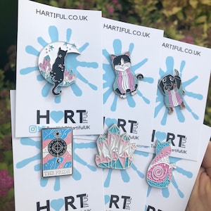 May include: A collection of enamel pins featuring various designs. The pins include a black cat on a crescent moon, a cat in a sweater, a dachshund in a sweater, a tarot card with the text "THE PRIDE", a crystal, and a cat with a spiral design. The website HARTIFUL.CO.UK is visible.
