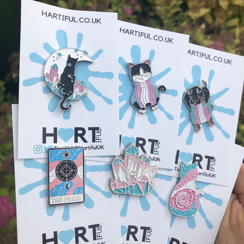 May include: A collection of enamel pins featuring various designs. The pins include a black cat on a crescent moon, a cat in a sweater, a dachshund in a sweater, a tarot card with the text "THE PRIDE", a crystal, and a cat with a spiral design. The website HARTIFUL.CO.UK is visible.
